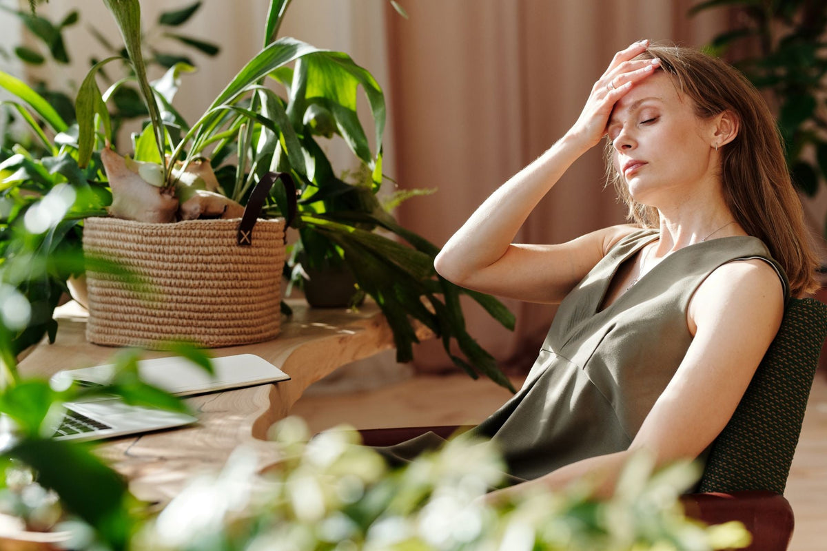 Woman with hand on forehead sitting indoors among plants, showing stress or fatigue.